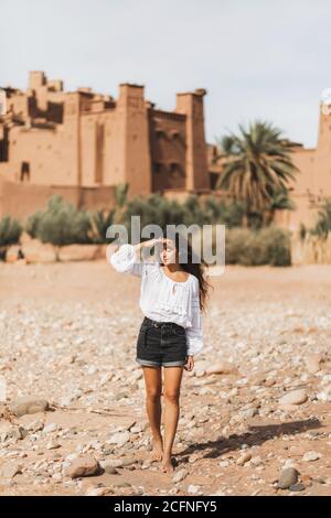 Young curly woman eastern appearance walking on background of kasbah Ait-Ben-Haddou. Travel in Morocco, Ouarzazate. Summer vacations, travel lifestyle concept. Stock Photo