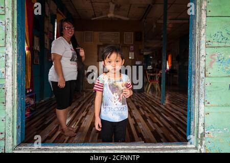 Bandar Seri Begawan / Brunei - January 16, 2019: portrait of little shy pretty girl looking at the camera at the door of wooden house in Kampong Ayer floating village with her mother next to her Stock Photo