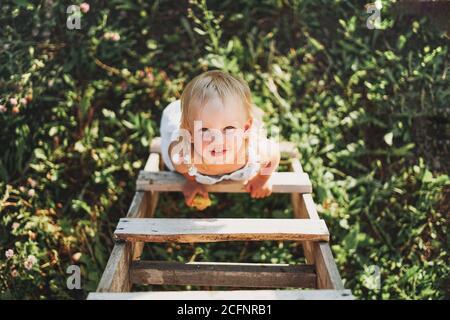 Cute blonde toddler little girl climbs up the ladder, country side, cottagecor Stock Photo