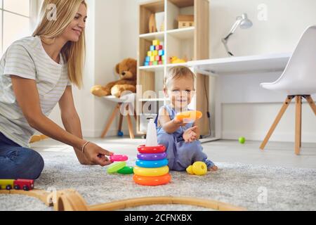 Happy young mother and her little son together assemble a colorful pyramid sitting on the floor.