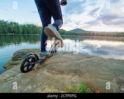 Blue sports wearing boy stay with fun push scooter on cliff at lake and makes tricks. Evening sun at horizon makes flares. Stock Photo