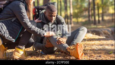 Unrecognizable woman comforting injured black guy, backpacking together by forest Stock Photo