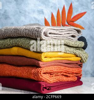 Stack of folded warm knitted women's sweaters in warm colors and bright autumn leaves on gray background. Copy space. Closeup Stock Photo