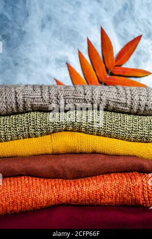 Stack of folded warm knitted women's sweaters in warm colors and bright autumn leaves on gray background. Copy space. Closeup Stock Photo