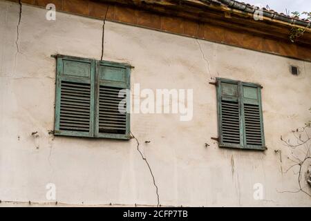 Facade of an abandoned building with old windows with green shutters. Plastered weathered wall Stock Photo