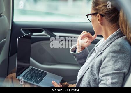Working on the way to the office, side view of a thouthfull caucasian business woman working on laptop while sitting on back seat in the car Stock Photo