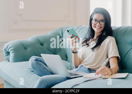 Pleased young female designer with dark hair creats new project, holds modern smartphone in hands, makes notes in notepad, poses on sofa, uses laptop Stock Photo