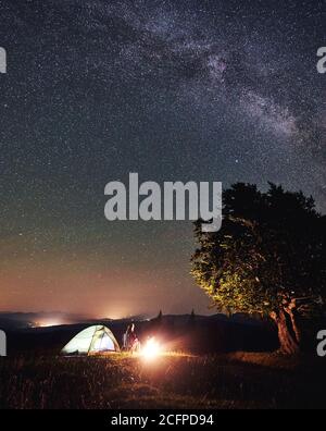 Female hiker relaxing at summer night camping in the mountains beside bonfire, illuminated tourist tent and big tree. Woman sitting on chair, enjoying view of night sky full of stars and Milky way. Stock Photo