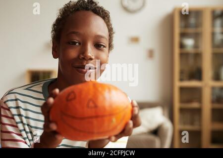 Medium close-up portrait of joyful African American kid holding ripe pumpkin with jack-o'-lantern face drawn on it, copy space Stock Photo