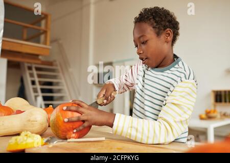 Portrait of preteen African American boy wearing casual outfit standing at table in kitchen carving pumpkin for Halloween Stock Photo
