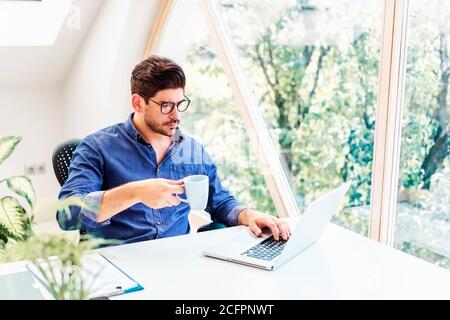 Shot of thinking businessman sitting at office desk and drinking morning coffee while working on laptop. Home office. Stock Photo
