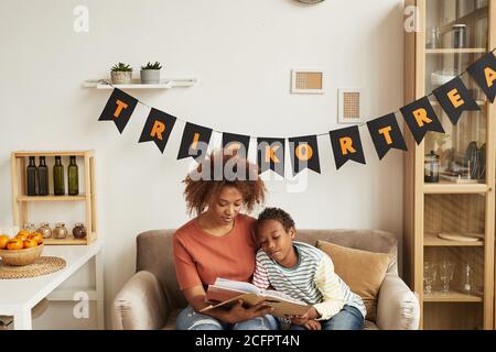 Modern African American mother sitting on sofa in living room with her young son reading spooky Halloween stories