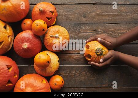 Directly from above view flat lay shot of carved pumpkins and tangerines with jack o' lantern faces and womans hands holding one pumpkin for Halloween on dark wooden table background... Stock Photo