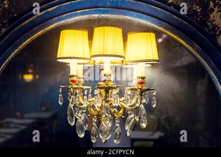 a candelabrum with a yellow lampshade with glass elements on an old mirror with a worn wooden frame, a wall lamp in a dark retro interior shines with Stock Photo