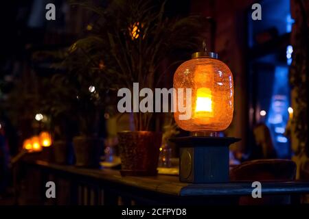orange glass lantern decorated with cracks installed on the wooden railing of the terrace of a city restaurant with flowerpots, night scene illuminati Stock Photo