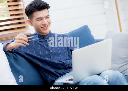 Young asian man sit on sofa using laptop computer shopping online with credit card buying to internet, happy male payment with business on couch, purc Stock Photo