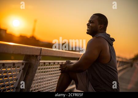 Young african-american man is having pain in knee while exercising in sunset on the bridge in the city. Stock Photo