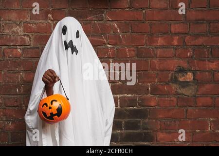Unrecognizable kid dressed up as ghost wearing white fabric holding jack o' lantern basket for sweets standing against brick wall Stock Photo