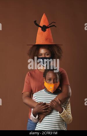 Vertical studio portrait of modern young woman wearing witch hat standing with her her cute son both wearing protective masks, brown background Stock Photo