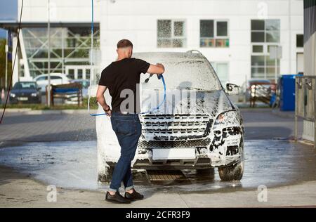 Back view of a sporty man holding a hose with soap and washing his car outdoors, white foam on a black car Stock Photo