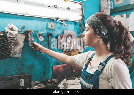 Gender equality. Portrait of a young smiling woman in uniform working in a workshop, who takes tools from a tool box on the wall. Stock Photo