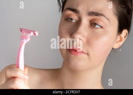 A young pretty woman with a dissatisfied expression is holding a razor in her hands.The concept of getting rid of unwanted hair Stock Photo