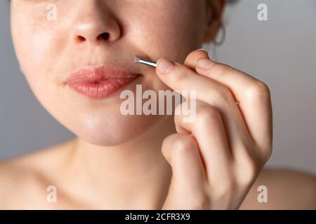 A young woman plucks her hair over her upper lip with tweezers. The concept of getting rid of unwanted facial hair. Close up. Stock Photo