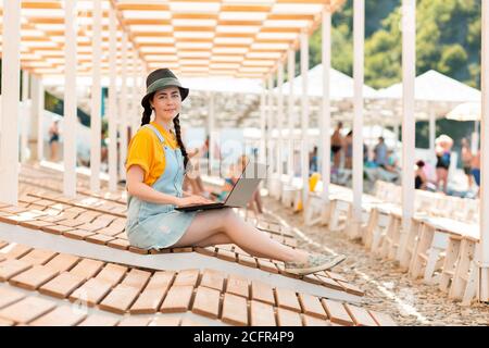 A young pretty woman is sitting on a chaise longue with a laptop and smiling. Beach umbrellas in the background. The concept of freelancing and remote Stock Photo