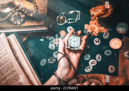 Astrology and esotericism. The female hand of the witch holds a watch on a chain. In the background, old books, fortune-telling runes, candles, and je Stock Photo