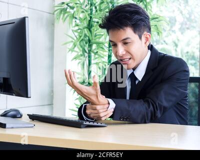 Young Asian businessman in suit getting hand pain while using notebook computer in office. Suffering from work-related pain and office syndrome concep Stock Photo