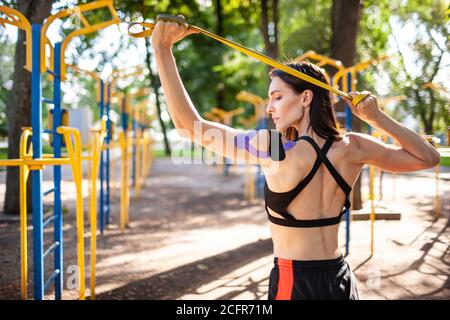 Brunette muscular woman posing with fitness resistance band in park, sports ground on background. Back view of young female with elastic taping on bod Stock Photo