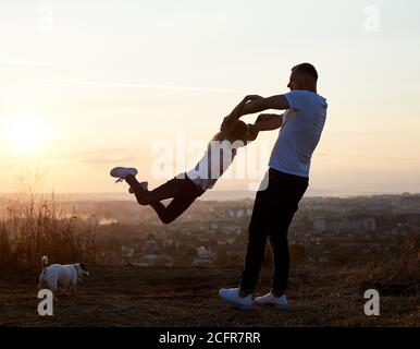 Silhouette of a father swinging his daughter by the arms on the hill on the setting sun in the suburbs, jack russell terrier near them, beautiful horizon on the background, side view