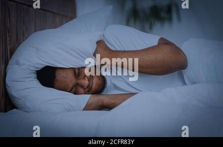 African Man Covering Ears With Pillow Having Insomnia At Home Stock Photo