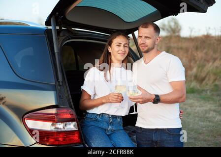 Portrait of a beautiful couple having some white wine near their black car, woman is sitting in the trunk embracing her husband, man is standing next to her, clinking glasses Stock Photo