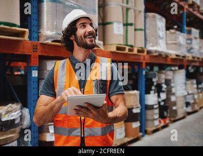 Waist up portrait of young man wearing reflective jacket holding digital tablet standing in factory warehouse smiling Stock Photo
