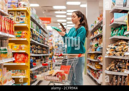 A young beautiful Caucasian woman reads the information on the label using her mobile phone. The concept of purchasing products and mobile apps. Stock Photo