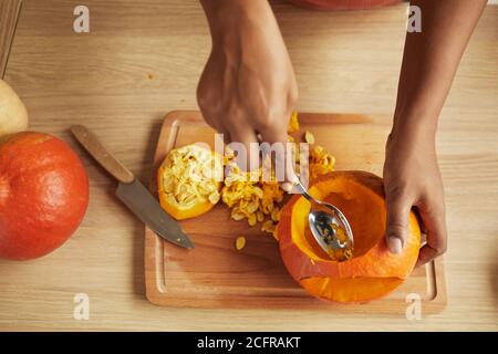 Top-down shot of womans hands scooping seeds and fibres out of small ripe pumpkin using spoon Stock Photo