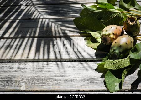 prickly pears nicely arranged on a white shabby chic wooden table with fig leaves , on the background a shadow of a palm, flat lay Stock Photo