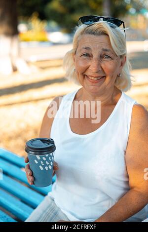 Adult blonde smiling woman with cup of takeaway coffee sitting on wood bench in green park on summer day, Stock Photo