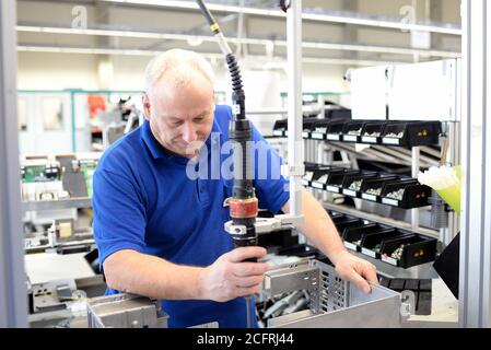 older friendly worker in an industrial company assembles electronic components in the mechanical engineering of a modern factory Stock Photo