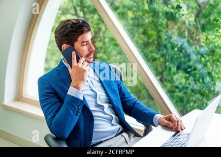 Shot of thinking businessman talking with somebody on his mobile phone while sitting at office desk and working on laptop. Home office. Stock Photo