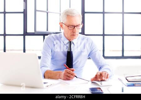 Portrait Of Senior Accountant Calculating Finance At Office. The businessman wearing shirt and tie and eyeglasses. The mature man working with laptop Stock Photo
