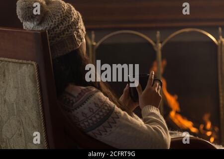 young Latin woman sitting on backwards, wearing a cap and wool sweater, in front of a lit fireplace, with a black cup in her hands Stock Photo