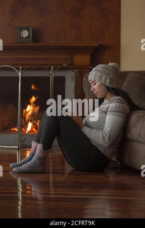 Young Latin woman smiling sitting on wooden floor with cell phone in hand, wears a woolen hat, colored jacket and black pants in the background a lit Stock Photo