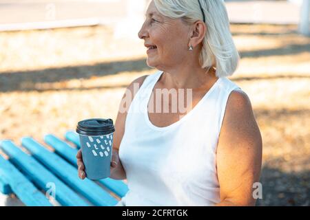 Adult blonde smiling woman with cup of takeaway coffee sitting on wood bench in green park on summer day Stock Photo