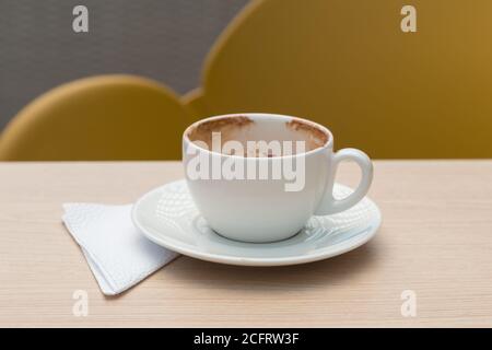 small white cup with chocolate, a plate and napkin, on a flat wooden surface, with a yellow chair in the background Stock Photo