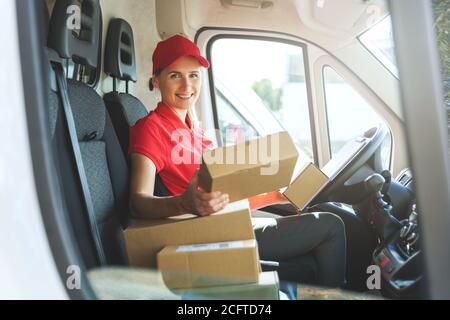 female delivery service worker in red uniform sitting in van with boxes. smiling at camera Stock Photo