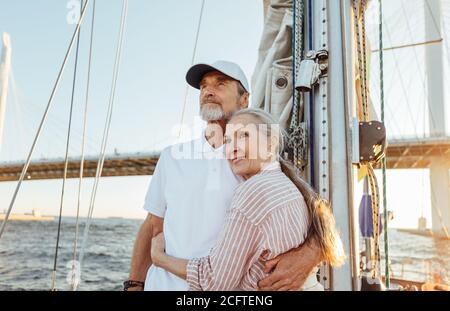 Affectionate senior couple hugging together and looking into the distance while standing on a yacht Stock Photo