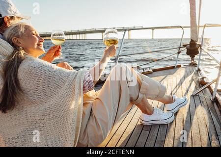 Mature man and woman wrapped in plaid on yacht deck and drinking wine. Senior couple holding glasses of wine on sailbot. Stock Photo