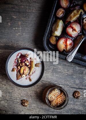 Roasted plums, red greengages and apples on baking pan. Buckwheat porridge with fruit and walnut in bowl. Honey on wooden table. Overhead view. Stock Photo